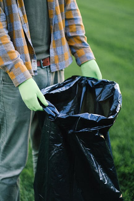 A person standing outdoors on a grassy surface, wearing a yellow, blue, and gray checkered shirt over a dark green top, with gray trousers secured by a brown belt, and wearing bright green gloves. The